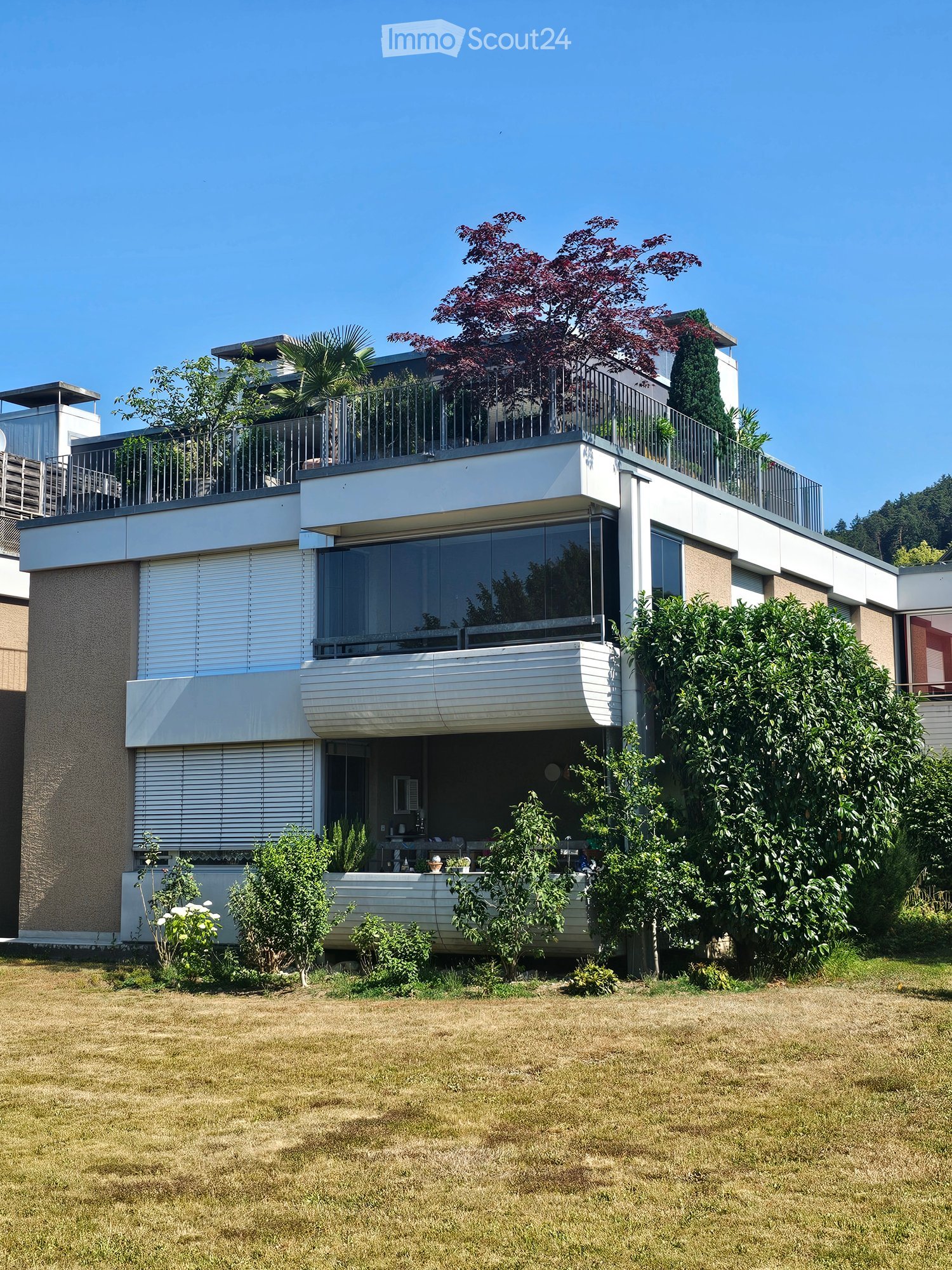 Modern two-story house with large windows, balconies, and surrounding greenery.
