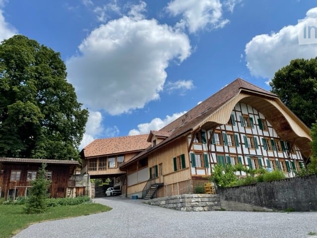 Traditional Swiss wooden house with green shutters, stone foundation, and a covered entrance. A small wooden outbuilding is visible to the left, and a car is parked in the driveway.