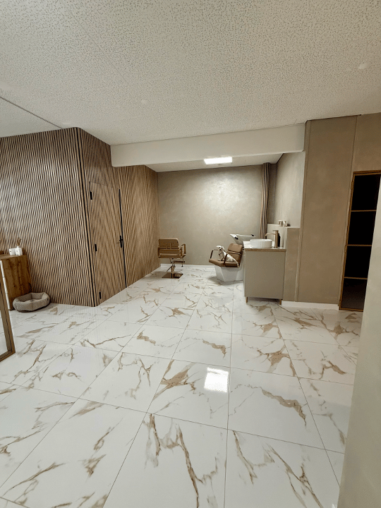 Modern, minimalist room with white marble flooring, wooden panel wall, and a sleek sink area. Two chairs and a stool are positioned in the center.