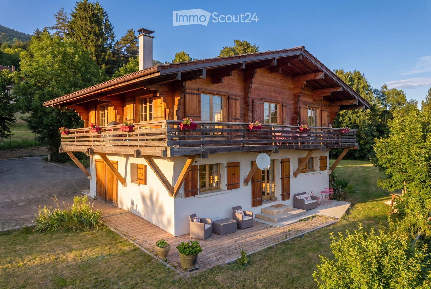 A two-story wooden house with a large balcony, surrounded by greenery and a well-maintained lawn. The house features a mix of wooden and white exterior walls, multiple windows with shutters, and a chimney. Outdoor seating includes a sofa, chairs, and a sm