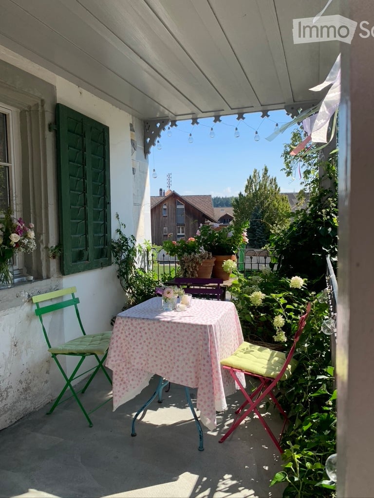 A cozy balcony with a small table and chairs, surrounded by lush greenery and flowers, with a view of the garden and neighboring houses.