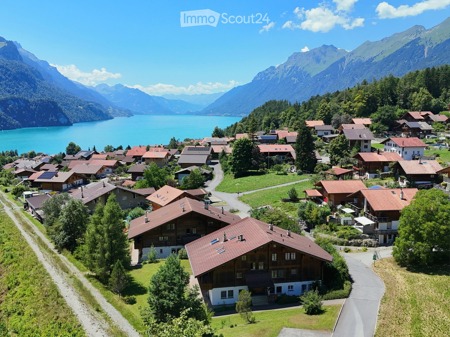 Aerial view of a village with traditional wooden houses, surrounded by lush greenery and mountains, with a lake in the background.