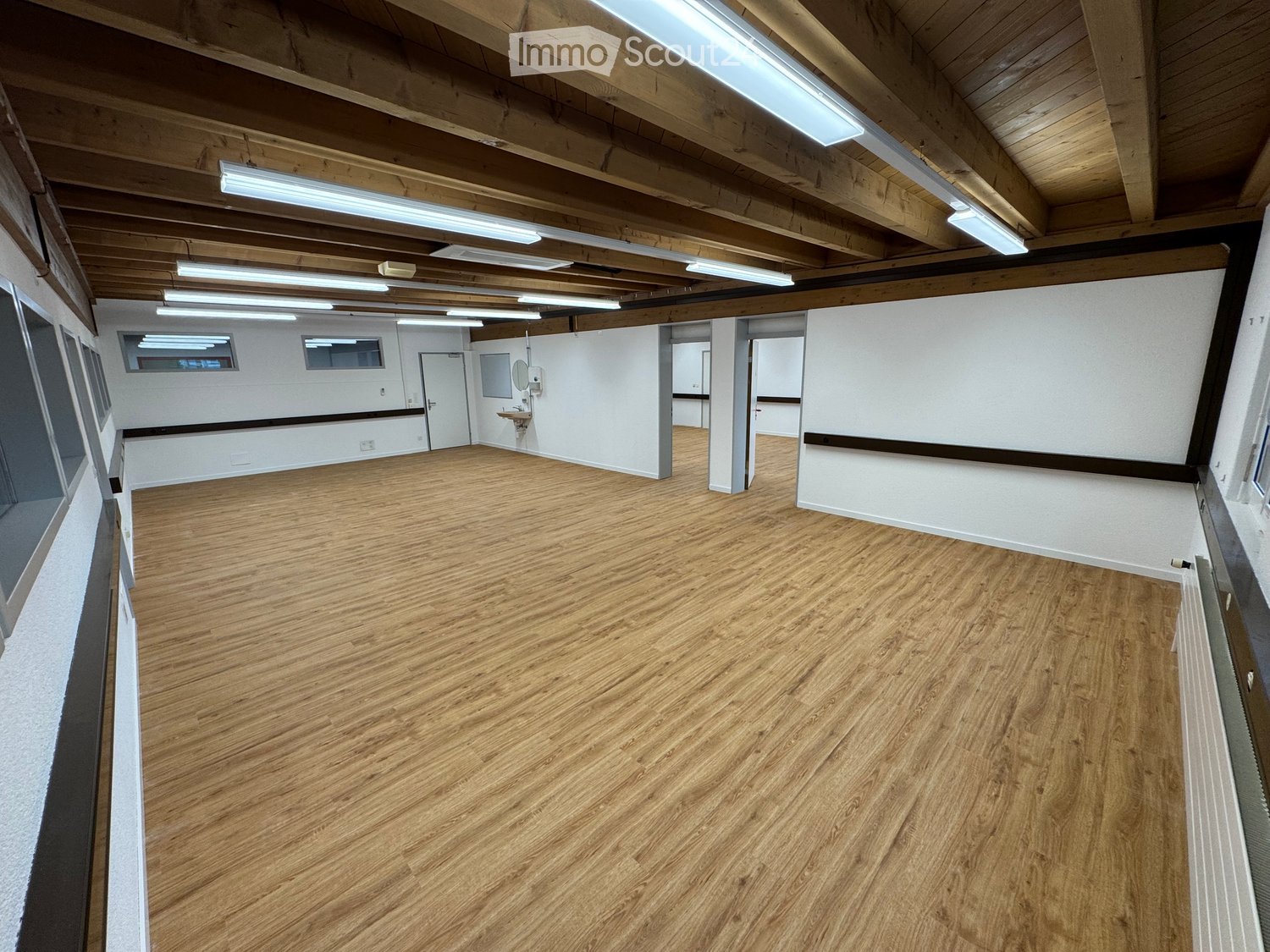 Empty room with wooden flooring, white walls, exposed wooden beams on the ceiling, and fluorescent lighting. Features a small bathroom area with a sink and mirror.