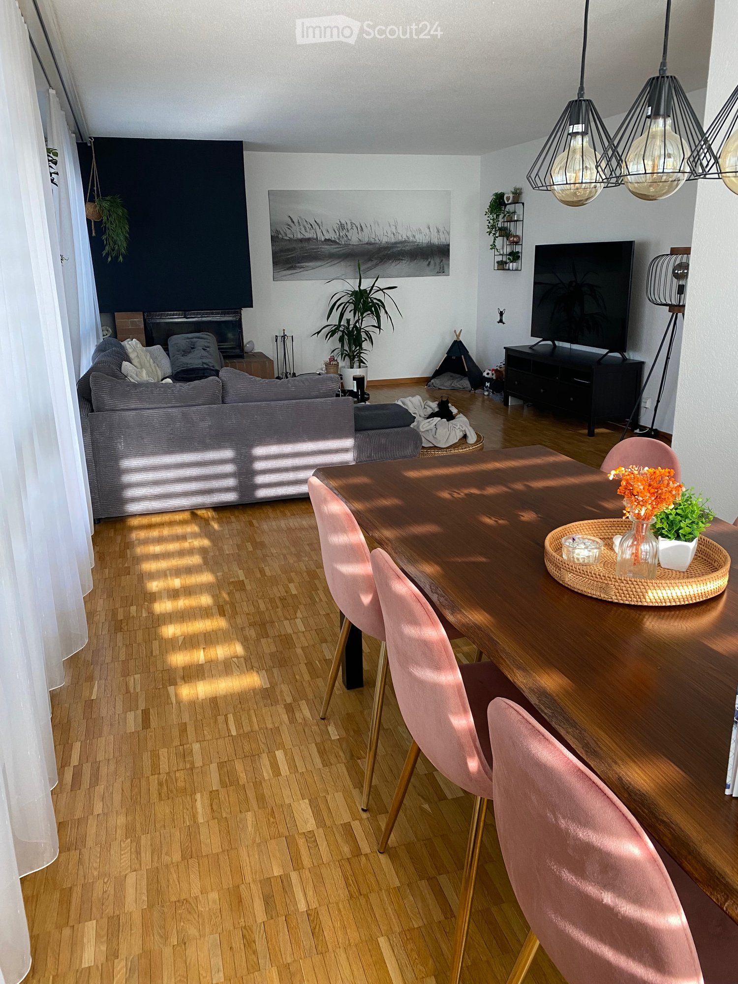 A modern living room with a grey sofa, wooden dining table, pink chairs, television, and decorative plants. Features a fireplace, artwork, and pendant lights.
