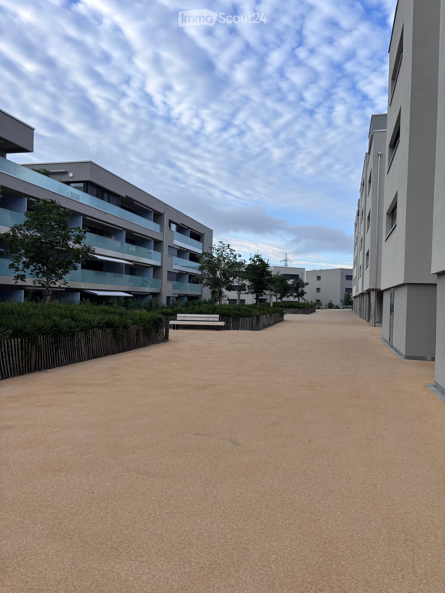 A wide, sandy pathway between modern apartment buildings with balconies, surrounded by greenery and a wooden bench.