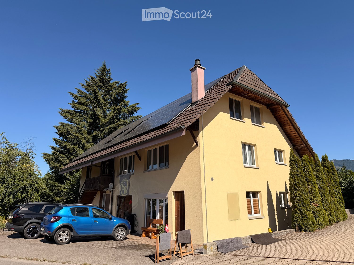 Two-story yellow house with a tiled roof, multiple windows, and a chimney. Solar panels are installed on the roof. There is a small outdoor seating area with chairs and a table near the entrance, and two parked cars in front of the house.