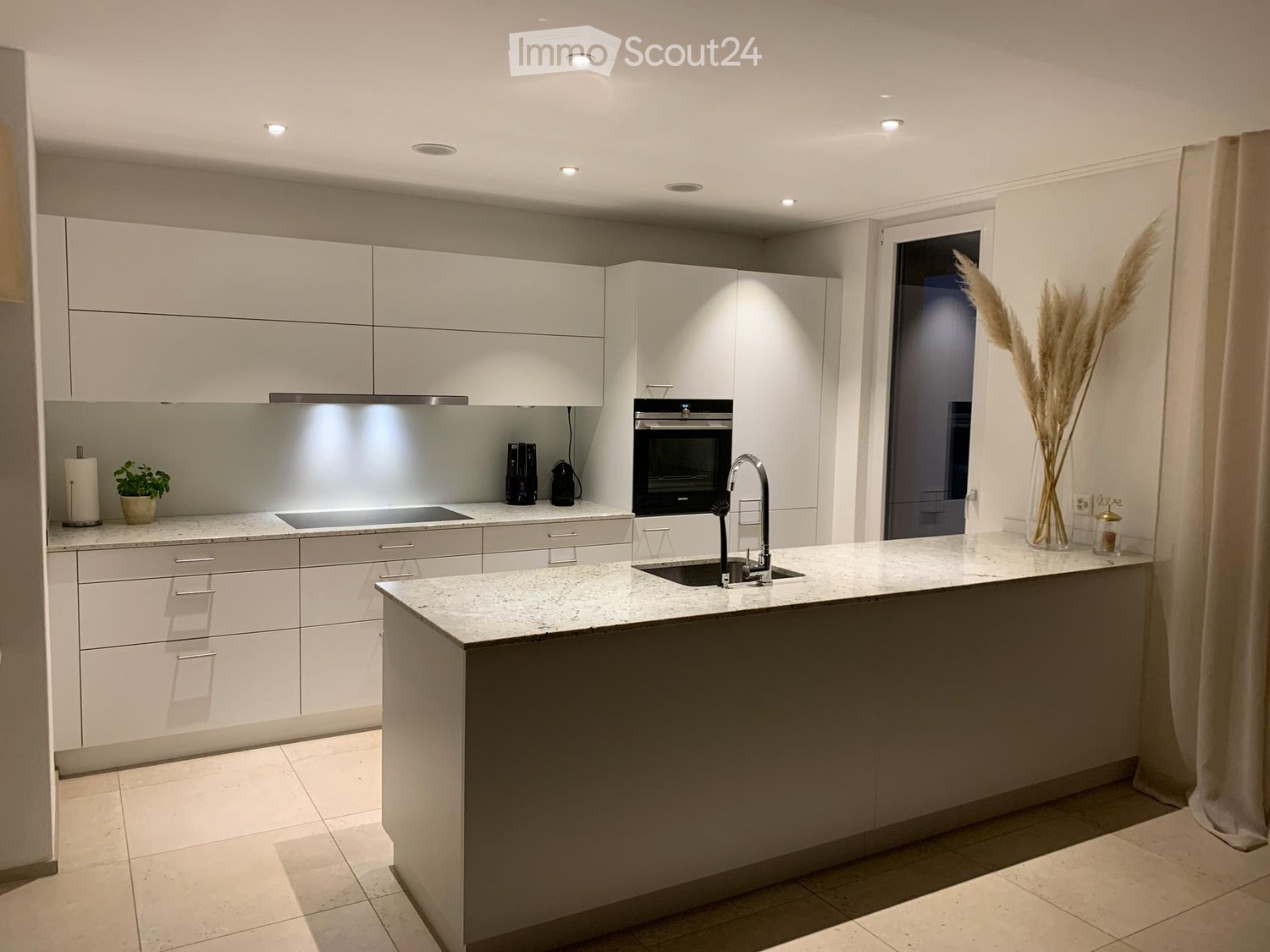 Modern kitchen with white cabinetry, a central island, and marble countertops. Features include a sink with a modern faucet, an oven, and decorative elements like a plant and a vase with dried flowers.
