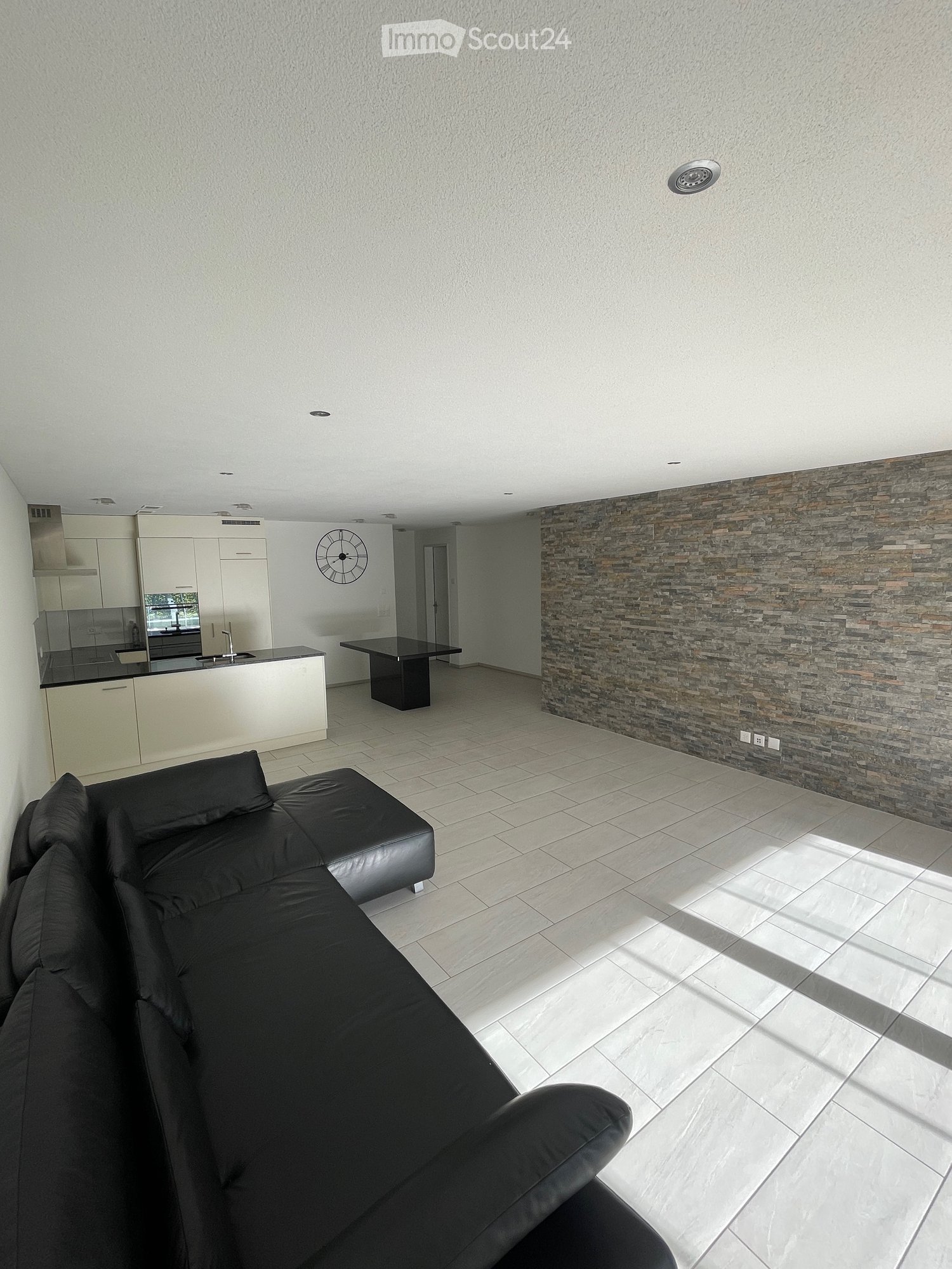 Modern living room with a black leather sofa, a black coffee table, a sleek kitchen area with white cabinets and a black countertop, and a stone accent wall.
