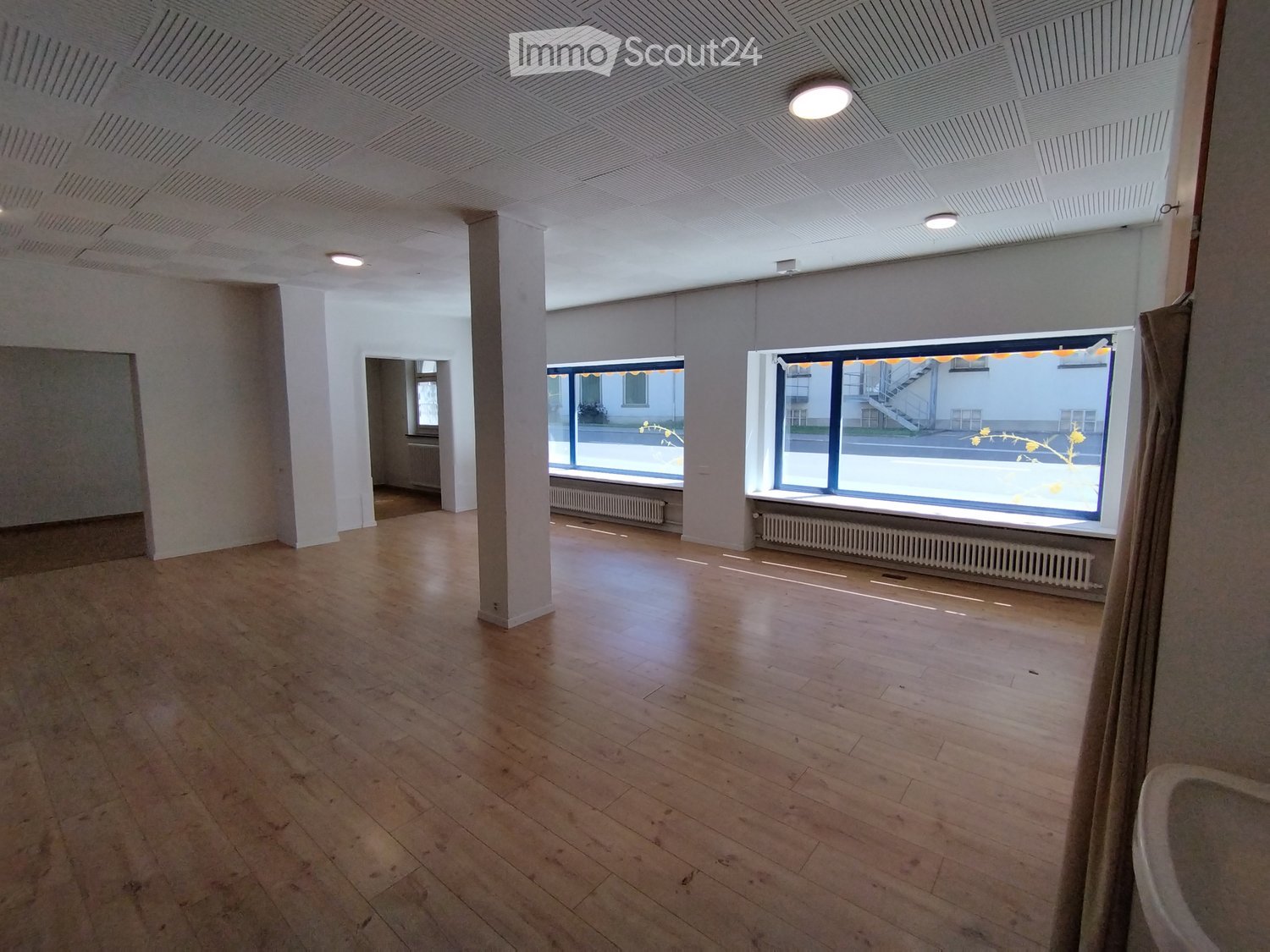 Empty room with wooden flooring, white walls, large windows, and a central column. No furniture or equipment present.