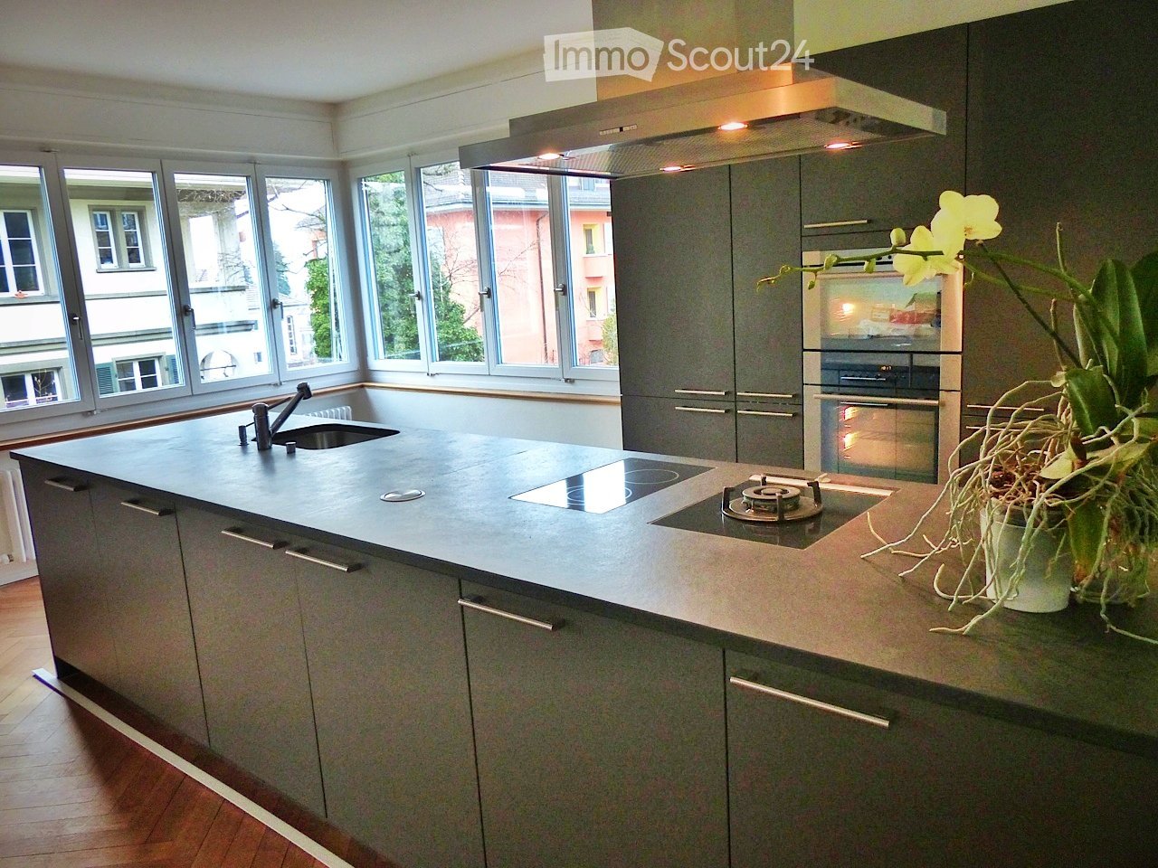 Modern kitchen with grey cabinets, a central island with a sink and stove, and large windows overlooking the street.