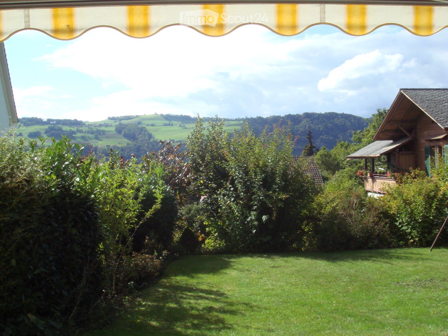 Lush green garden with well-maintained lawn, surrounded by trees and shrubs, with a scenic view of rolling hills and a wooden house in the background.