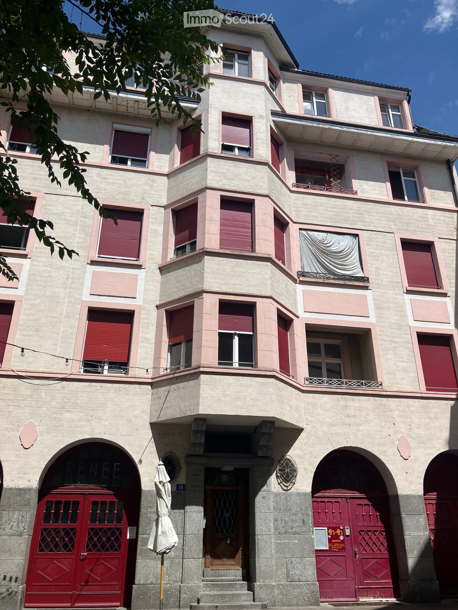 Four-story building with beige facade, red shutters, and arched red garage doors. Features a central entrance with a wooden door and umbrella stand.
