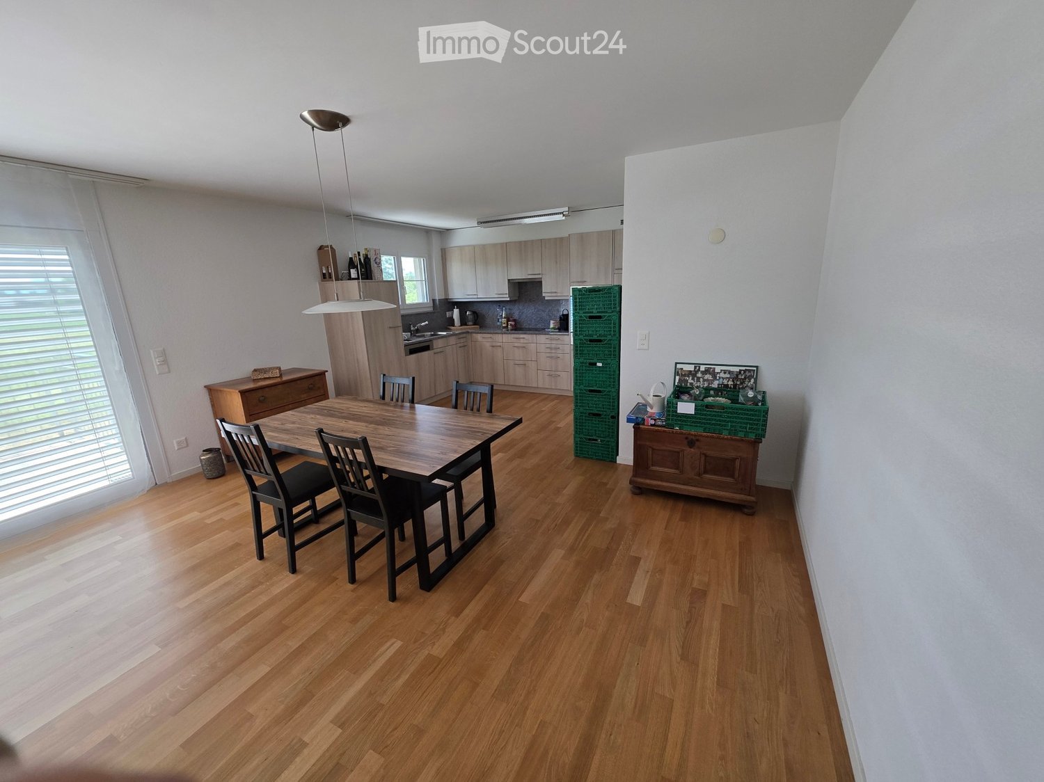 Bright, modern kitchen and dining area with light wooden flooring, white cabinets, and a central dining table with chairs. The kitchen features a sink, faucet, and various appliances, while the dining area includes a wooden table and chairs.