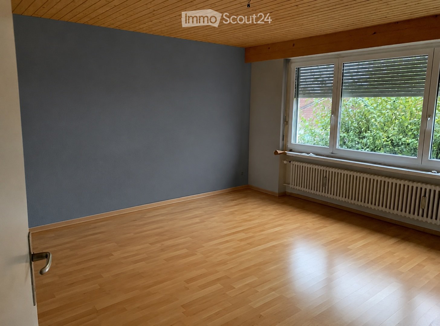 Empty room with wooden flooring, grey walls, large window with blinds, radiator, and a door with a silver handle.