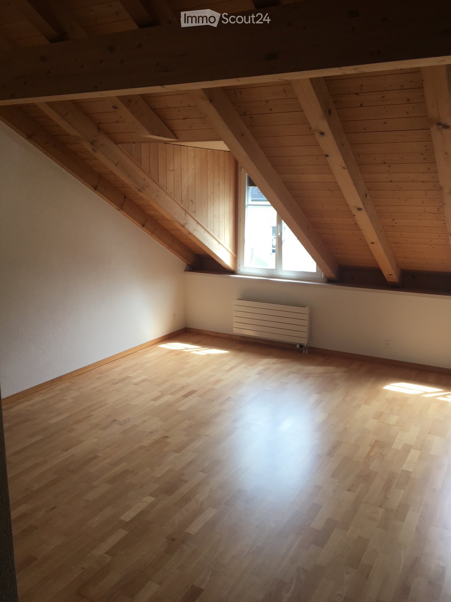 Empty attic room with wooden floor, exposed wooden beams, and a single window letting in natural light.