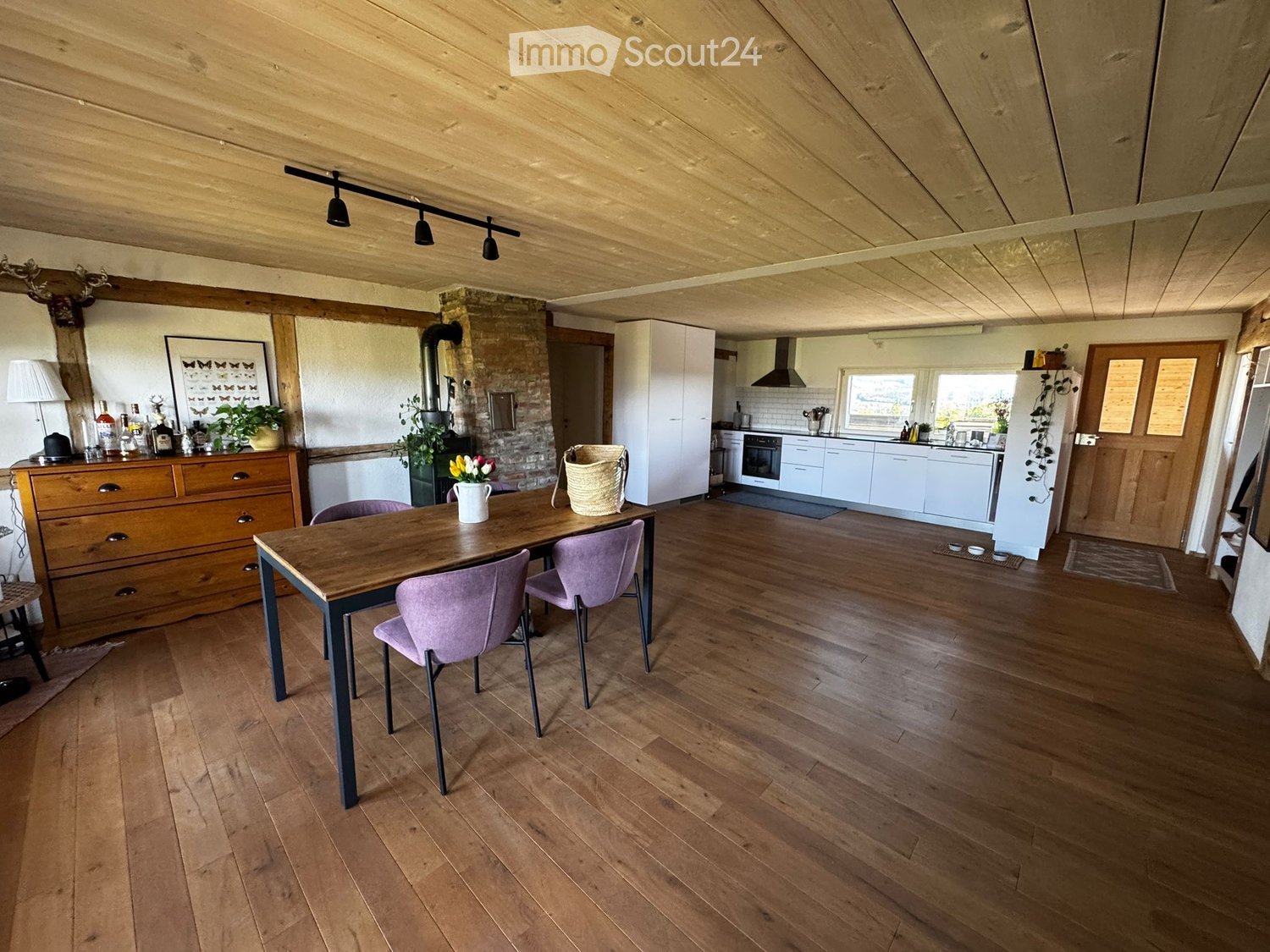 Open-plan living and dining area with wooden floors, exposed wooden beams, a dining table with chairs, a cabinet, and a modern kitchen area with white cabinets and stainless steel appliances.