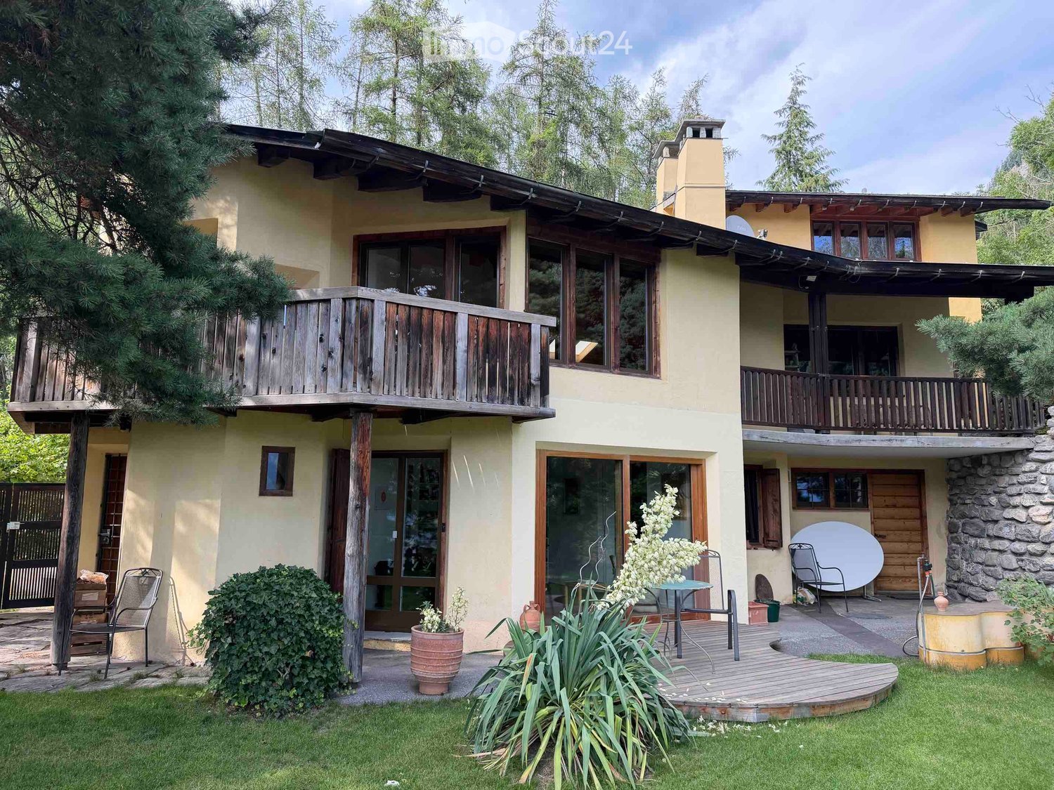 Two-story house with a wooden balcony, large windows, stone chimney, and outdoor seating area with a table and chairs.