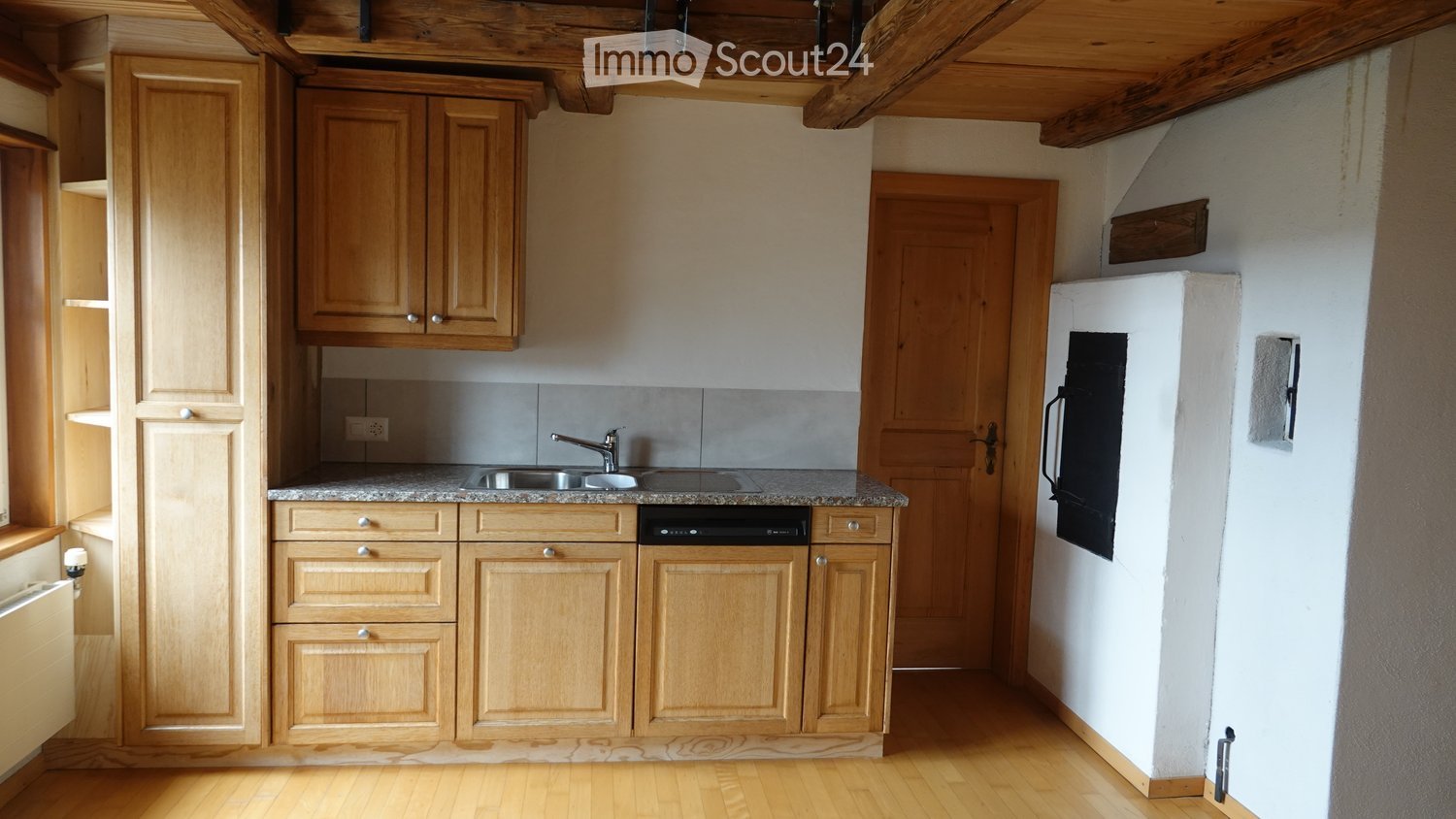Compact kitchen with light wooden cabinets, granite countertop, sink, dishwasher, and refrigerator. Wooden floor and exposed wooden beams on the ceiling.