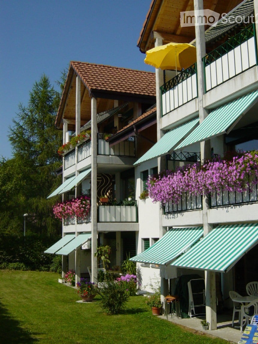 Multi-story residential building with balconies, green awnings, and flower boxes, surrounded by a well-maintained lawn and outdoor seating area.