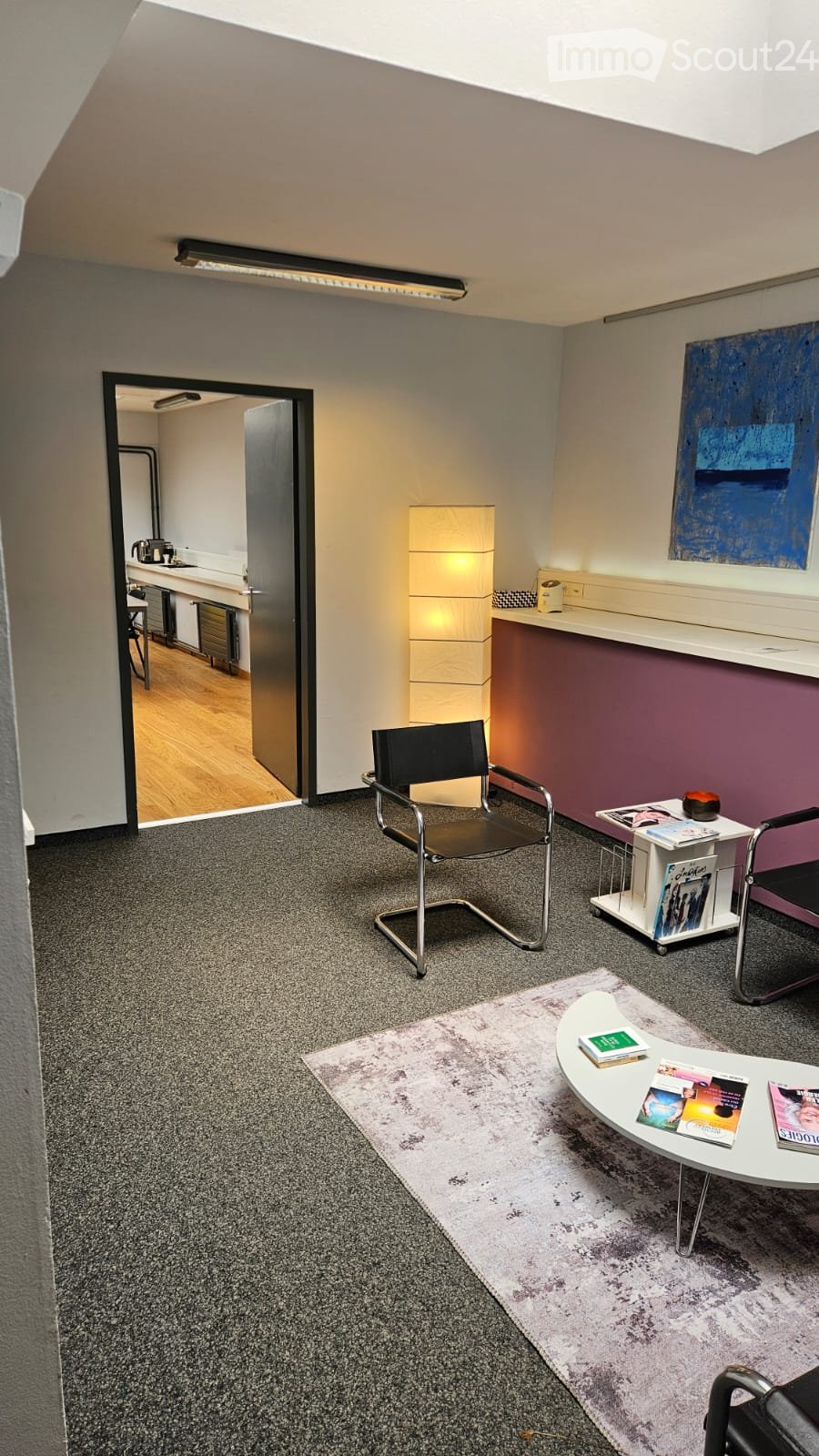 A modern, well-lit room with a grey carpet, white walls, and a slanted ceiling. Features include a sleek black chair, a white coffee table with books, a small side table with a lamp, and a colorful abstract painting on the wall. An open doorway leads to a