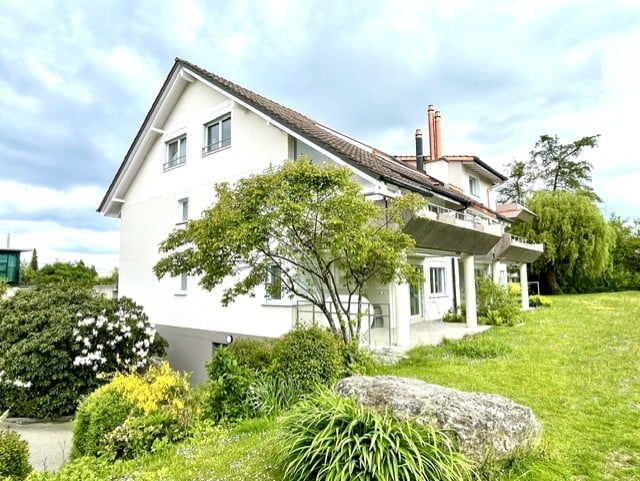 Two-story white house with a tiled roof, multiple windows, and a balcony overlooking a well-maintained garden.