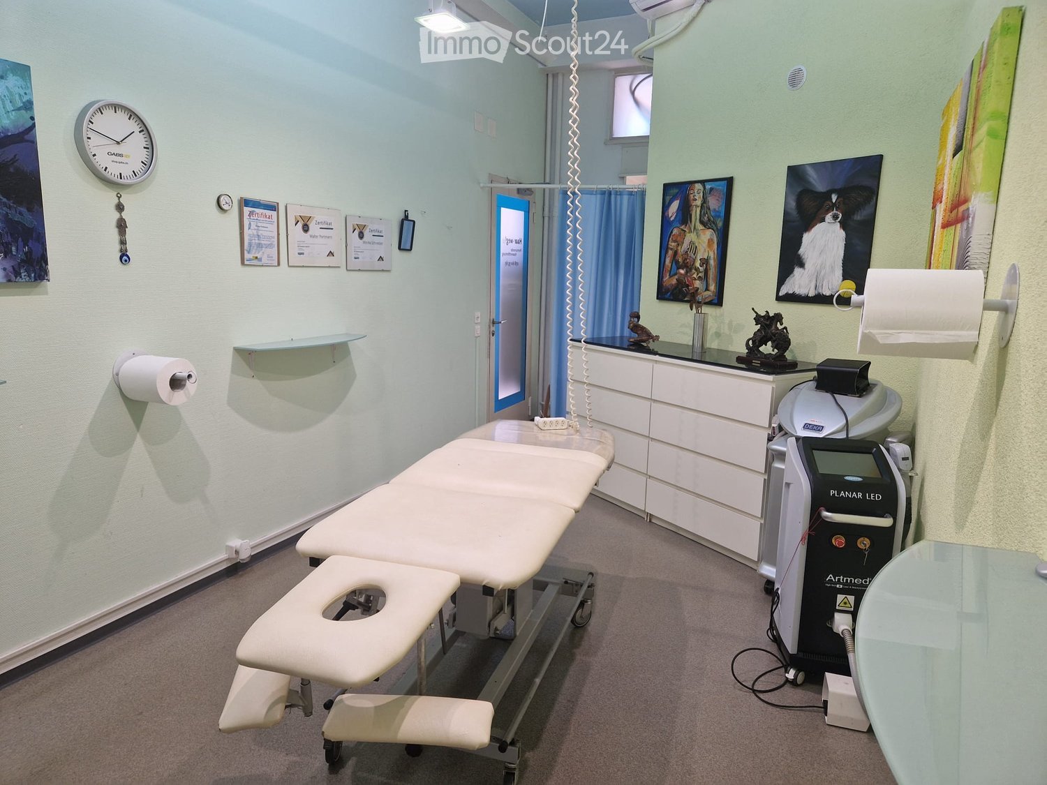 A clean, well-lit medical or treatment room with a treatment table, a chair, a cabinet, a medical device labeled "Artmed," a mirror, framed pictures, a clock, and toilet paper dispensers.