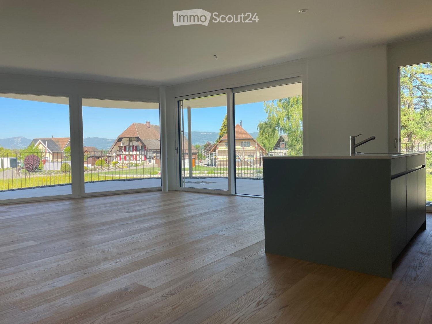 Empty room with wooden flooring, large sliding glass doors leading to a view of a suburban neighborhood, and a small kitchen counter with a faucet visible on the right side.