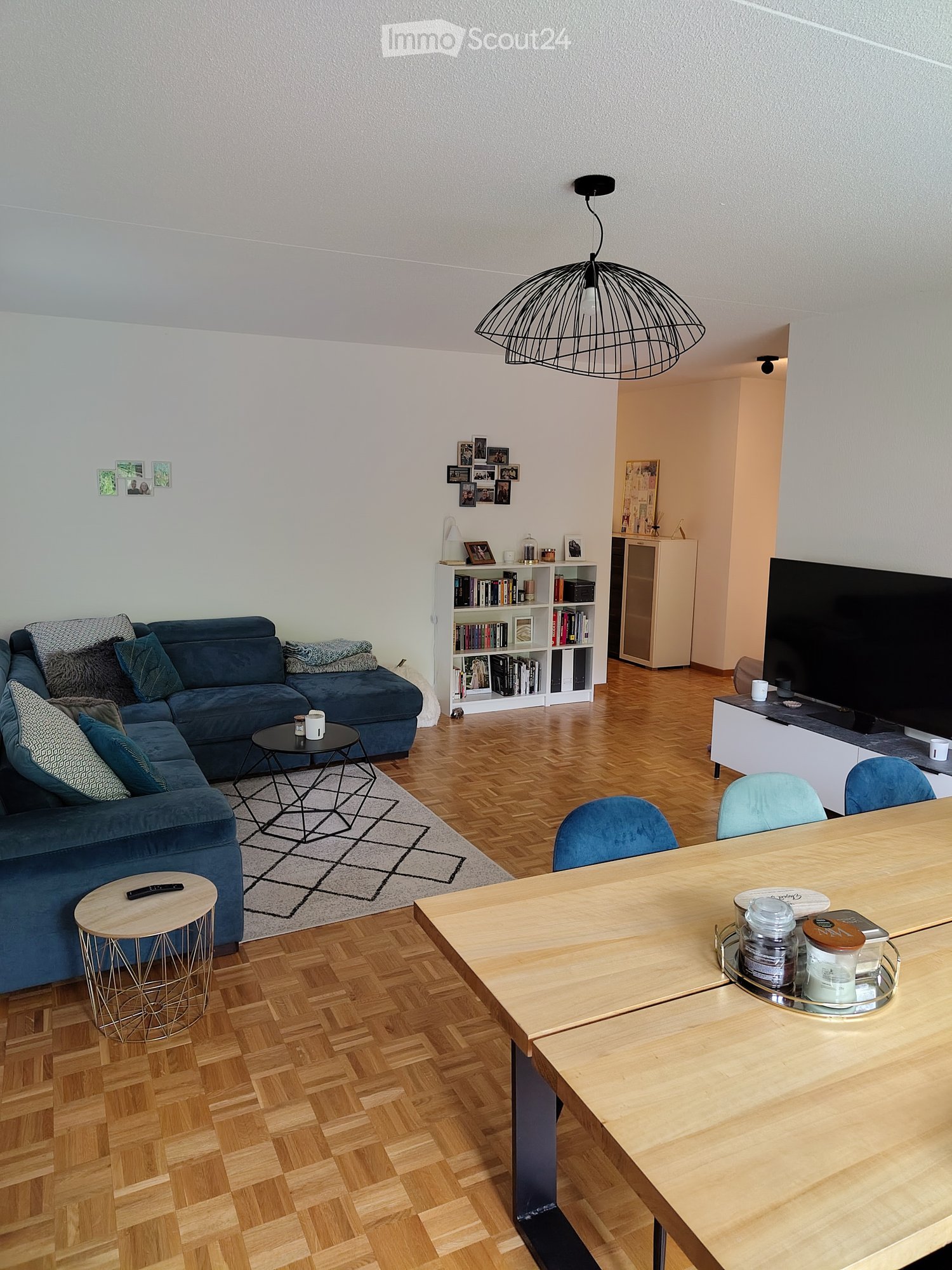 Modern living room with a blue sectional sofa, wooden coffee table, dining table with chairs, bookshelf, TV, and decorative jars.