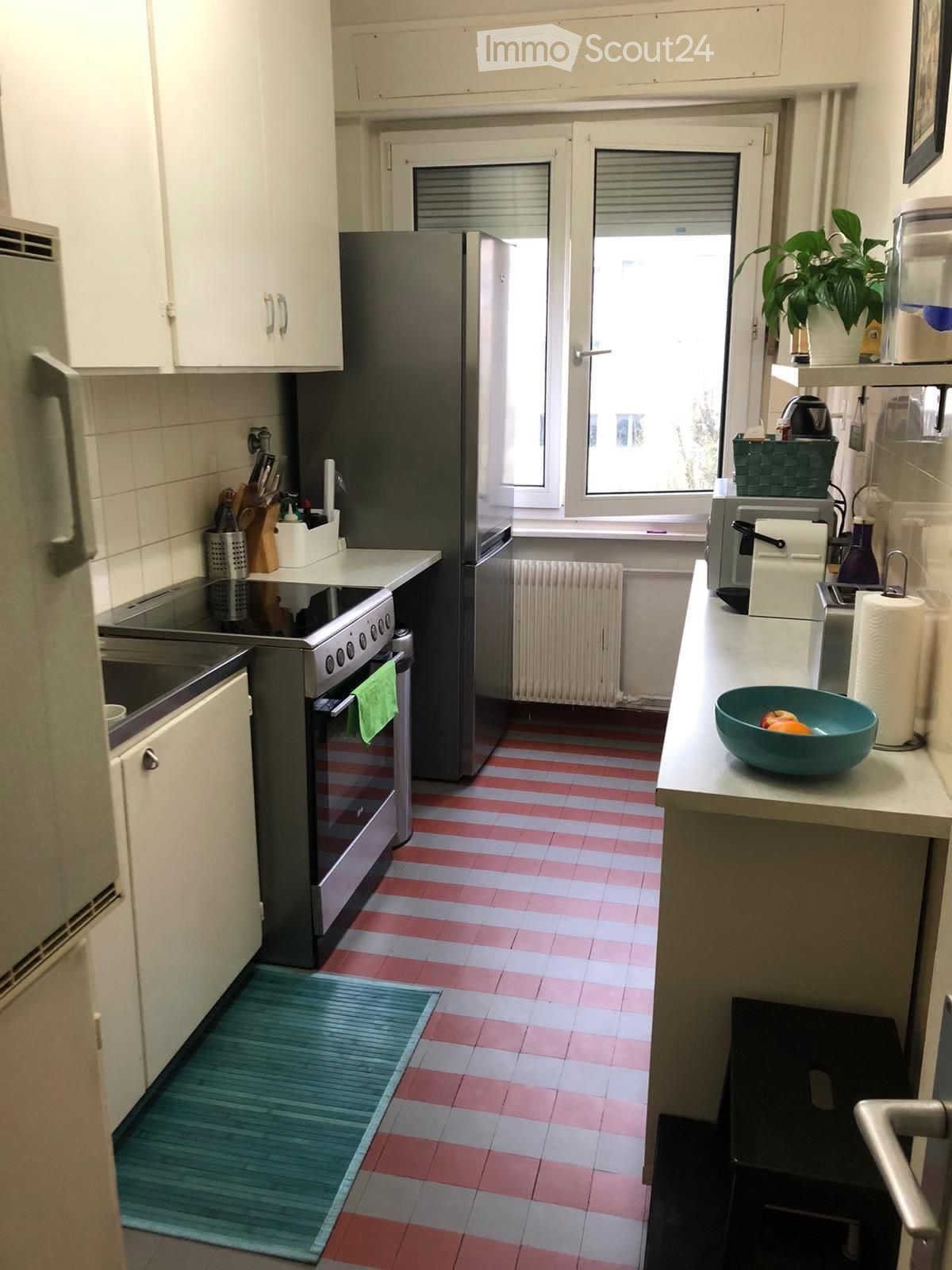 A compact kitchen with white cabinets, stainless steel appliances, and a striped red and white tiled floor. Features include a refrigerator, oven, sink, and various kitchen items on the counter.