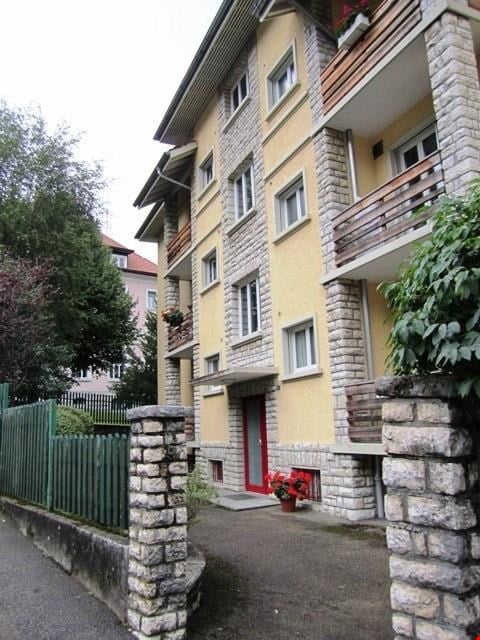 3 story yellow building, multiple balconies, stone pillar entrance, brick sidewalk, green fencing, red door