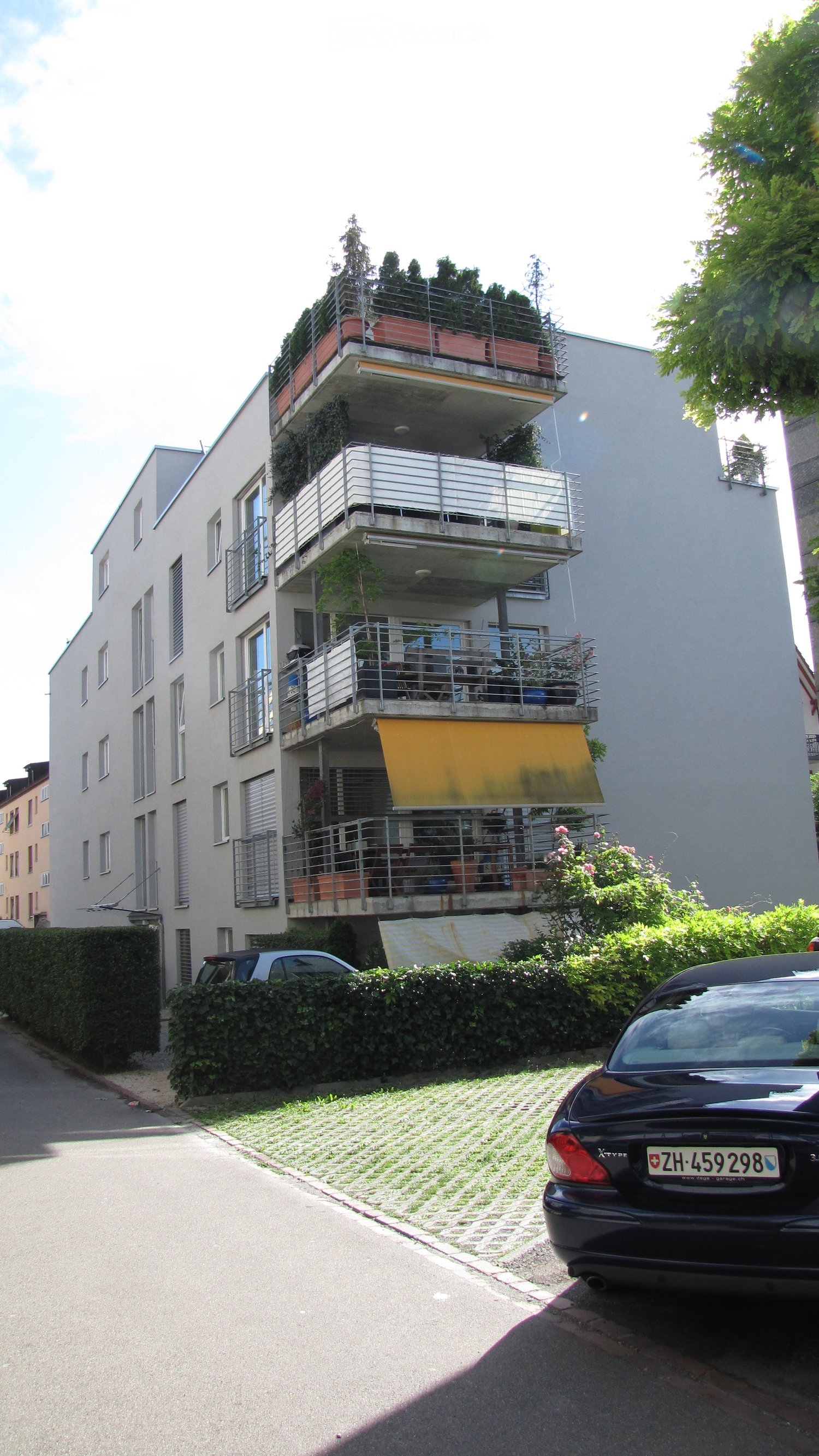 Multi-story residential building with multiple balconies, potted plants, and a parked car in front.