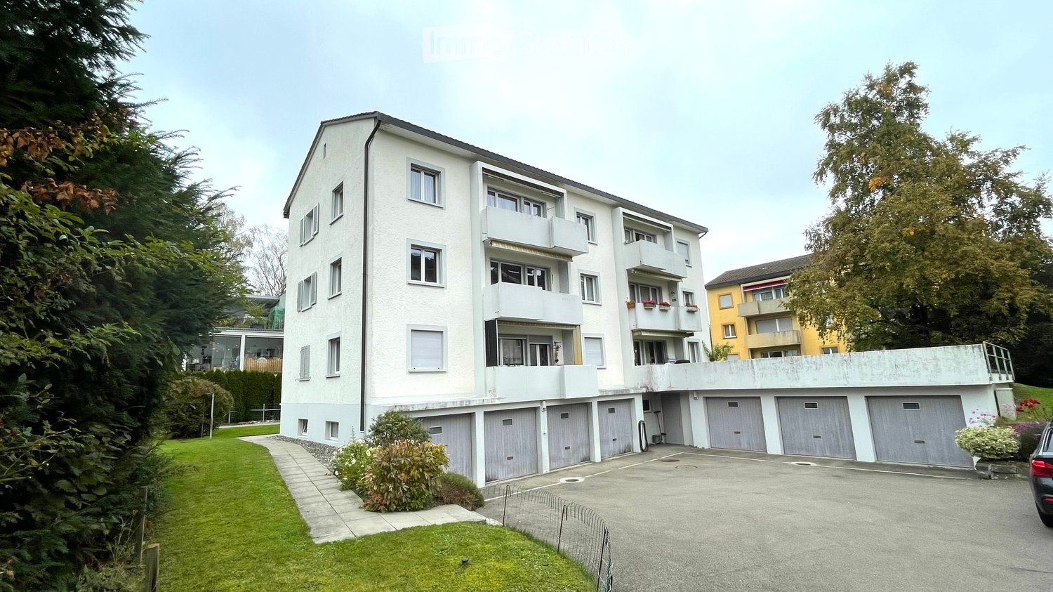 Modern multi-story apartment building with balconies, multiple garage doors, and surrounding greenery.