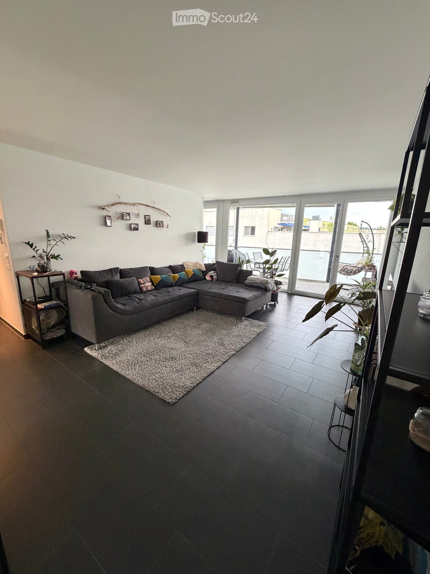 Modern living room with dark grey sectional sofa, colorful throw pillows, a white shag rug, dark tiled flooring, large sliding glass doors leading to a balcony, and decorative plants.