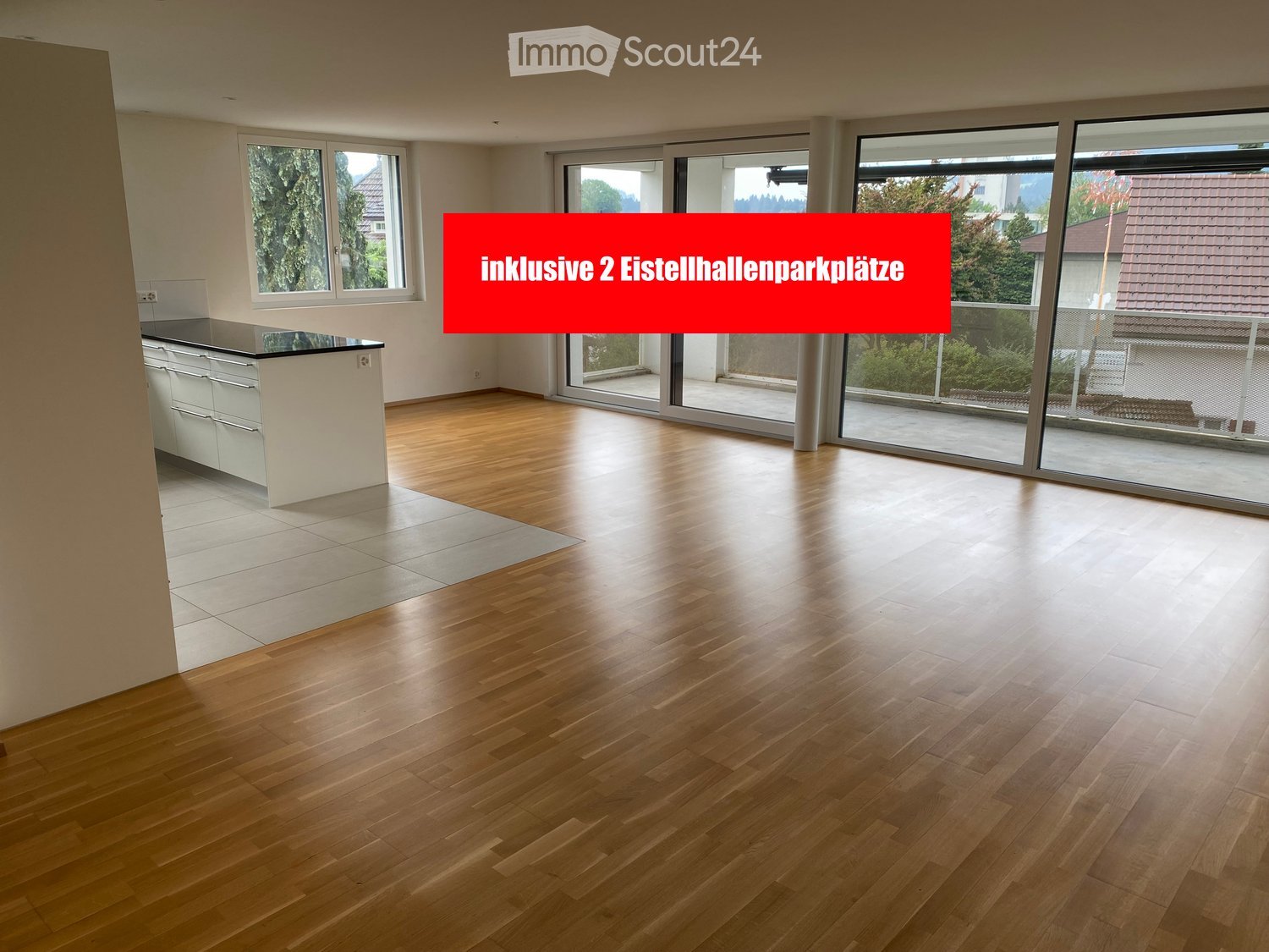 Empty room with wooden flooring, large windows, and a small kitchen area with cabinets and a countertop.