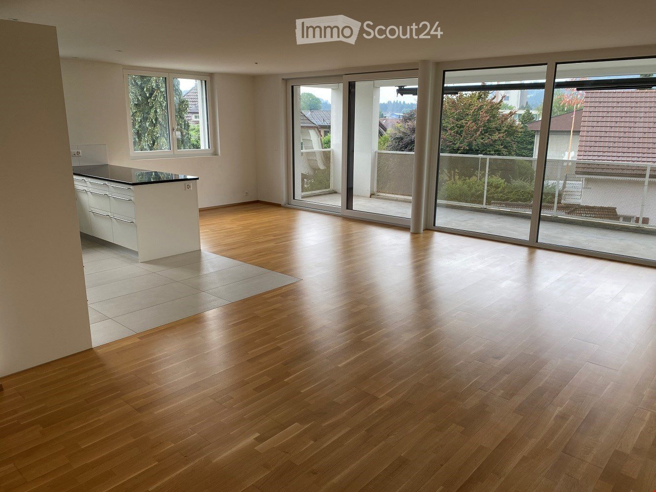 Empty room with wooden flooring, white walls, large windows, and sliding glass doors leading to a balcony.