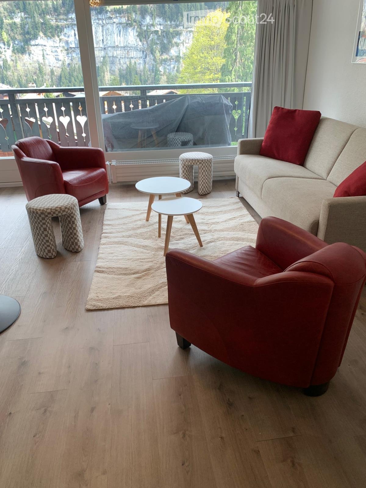 Modern living room with wooden flooring, large window offering a scenic view, a beige sofa with red pillows, red leather chairs, small white tables, and checkered stools.