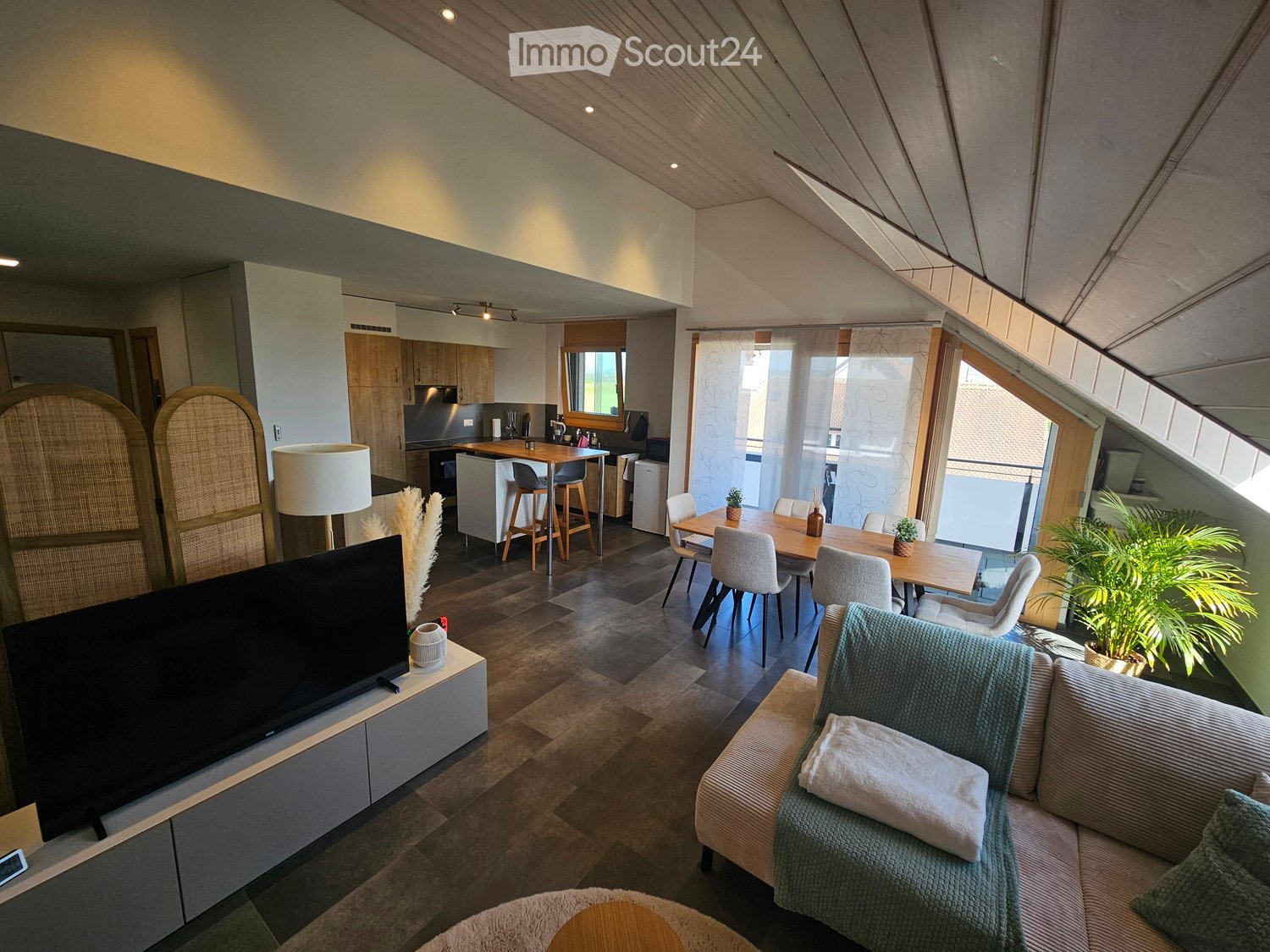 Open-plan living and dining area with a modern sofa, coffee table, TV, dining table with chairs, kitchen island, and bar stools. The space features wooden flooring, large windows, and a cozy, contemporary design.