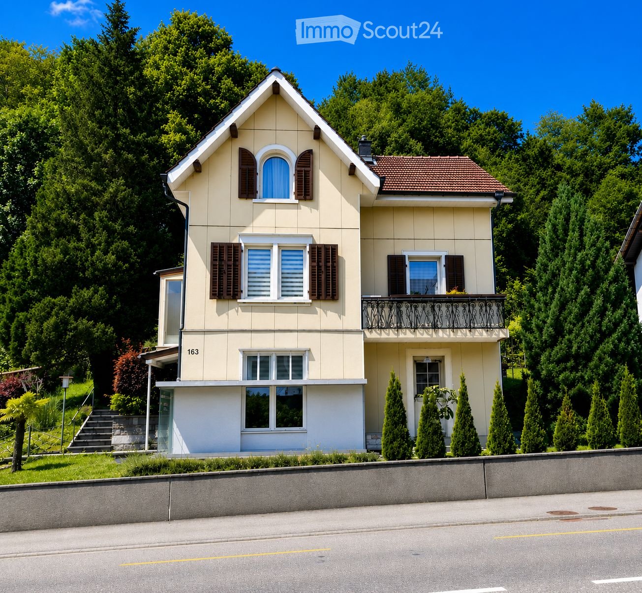 Two-story house with beige siding, multiple windows with shutters, a balcony on the upper level, and a small garden area.