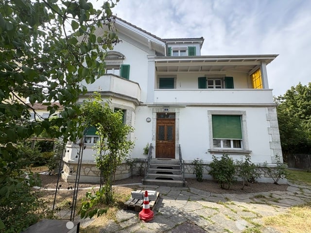 Two-story white house with green shutters, wooden front door, and a small balcony on the first floor.