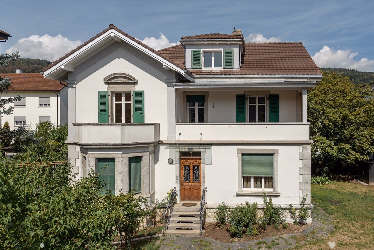 Two-story white house with green shutters, brown tiled roof, and a small balcony on the second floor. The entrance features a wooden door with decorative glass panels and a small set of steps leading up to it. The house is surrounded by greenery and trees
