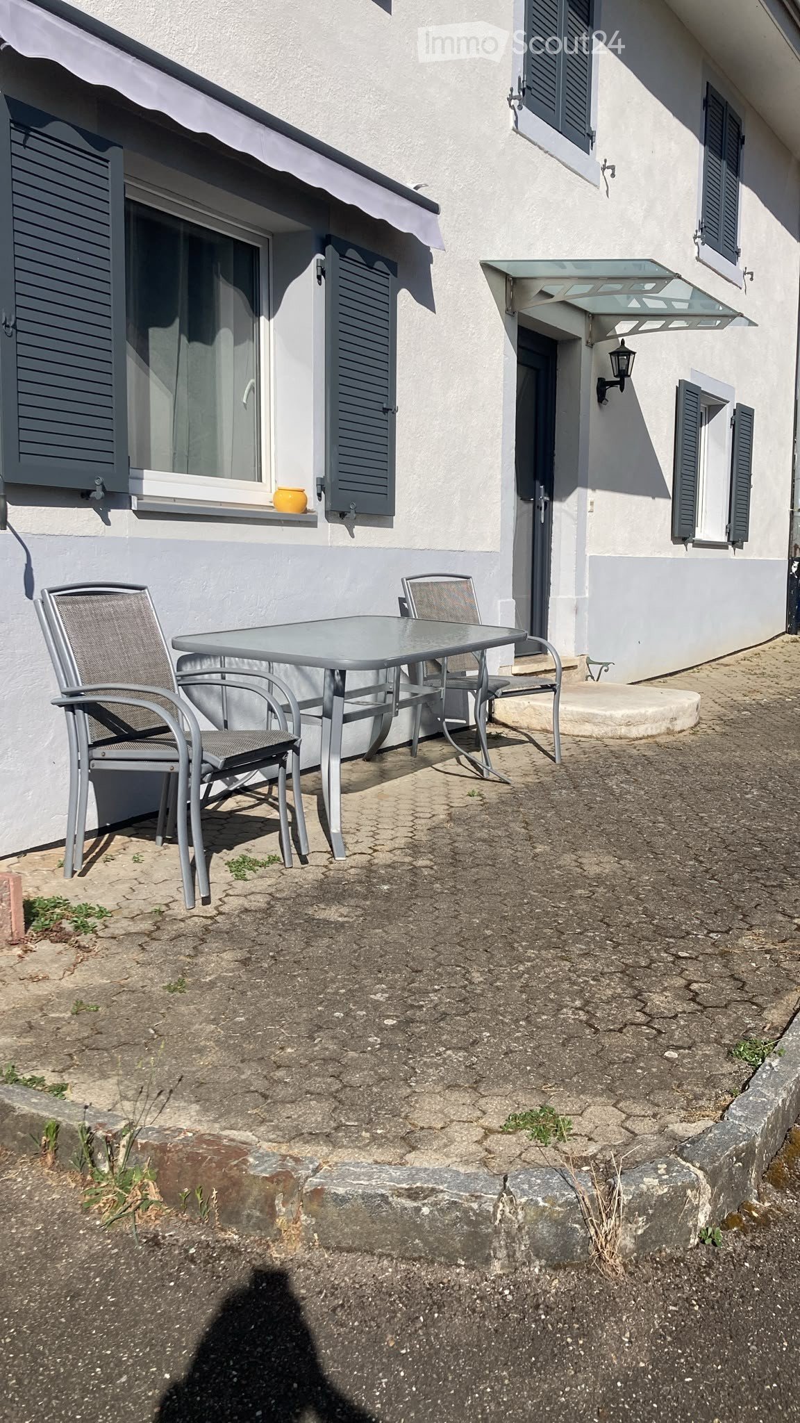 Outdoor seating area with a metal table and four chairs, situated next to a building with shutters and a small awning.