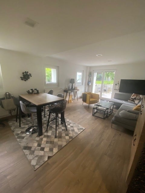 Bright, open-concept living and dining area with hardwood flooring, large windows, and a sliding glass door leading to a balcony. Features a grey sectional sofa, yellow armchair, coffee table, dining table with four chairs, side table with lamp, and wall