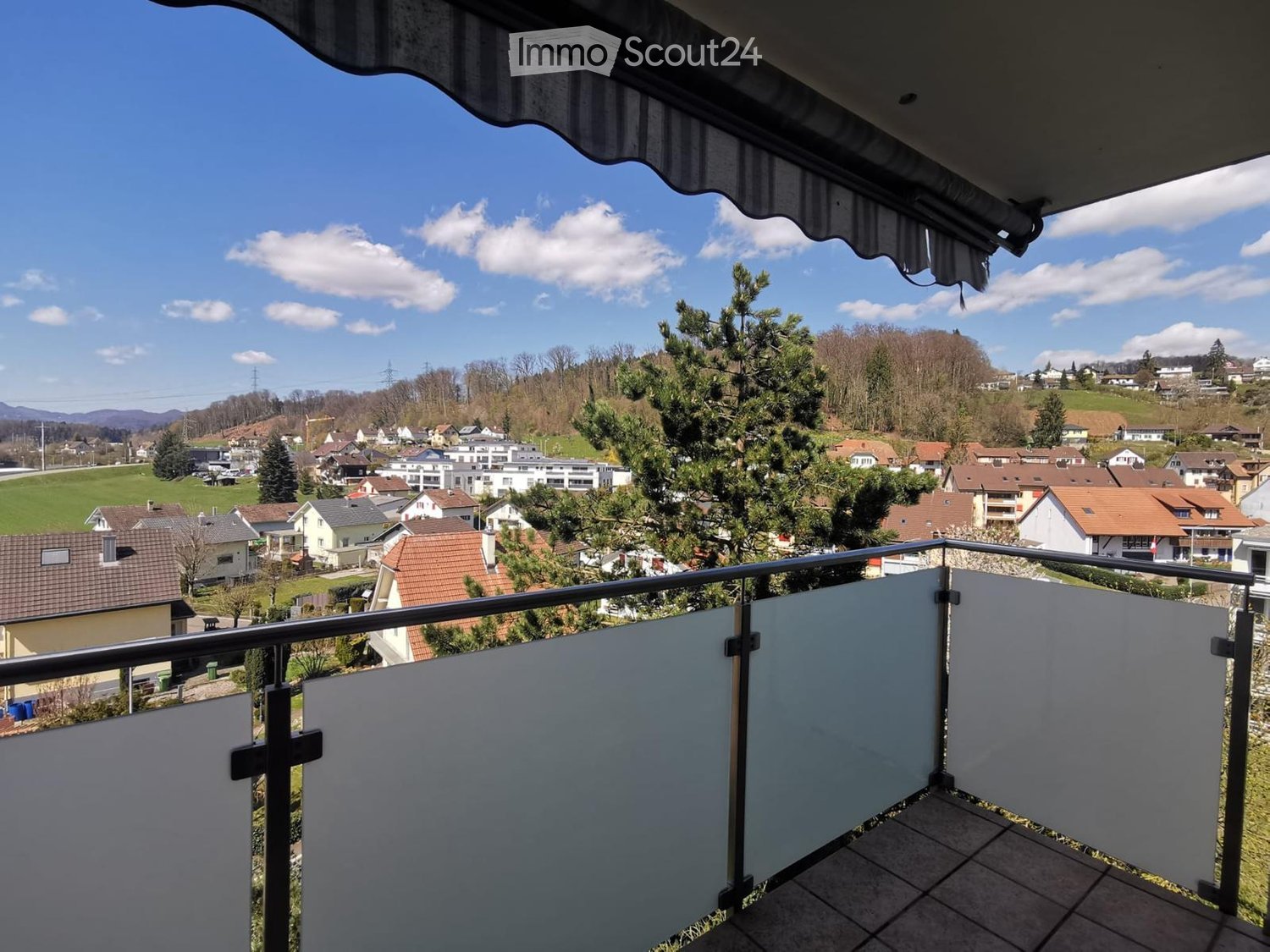 Balcony with metal railing and white privacy screens, overlooking a residential area with houses, trees, and a clear blue sky.