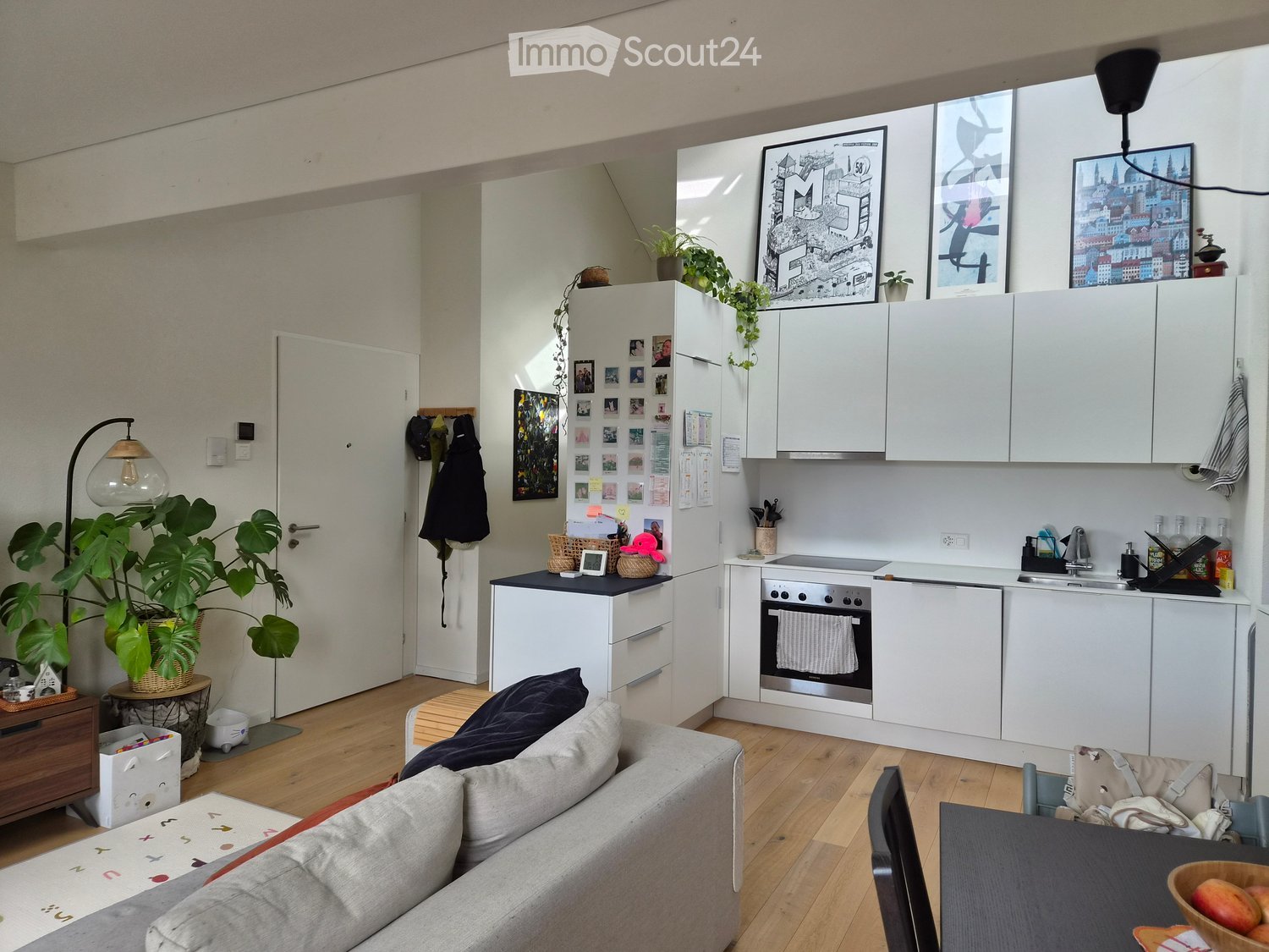 A modern, open-plan living and kitchen area with a grey sofa, a dining table with chairs, a fully equipped kitchen with white cabinets, an oven, and a sink. The space features wooden flooring, large windows, and decorative plants.