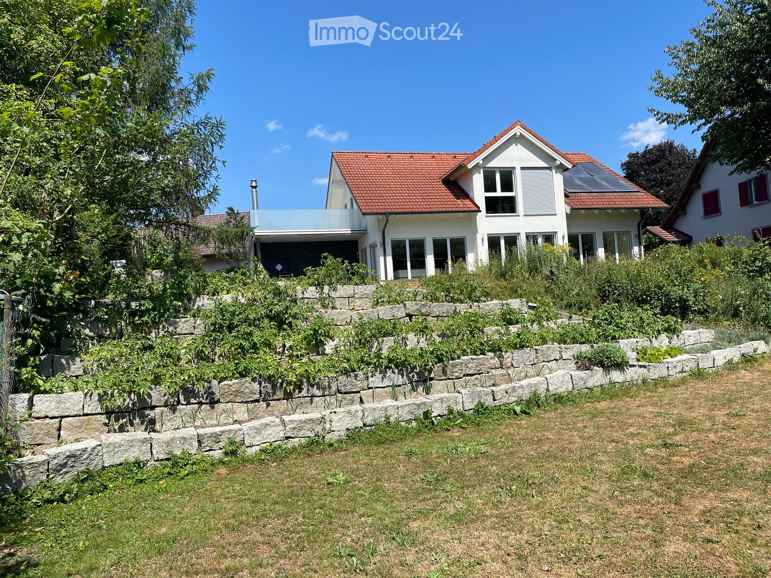 Two-story modern house with red-tiled roof, large windows, and a raised ground floor surrounded by a stone retaining wall and greenery.