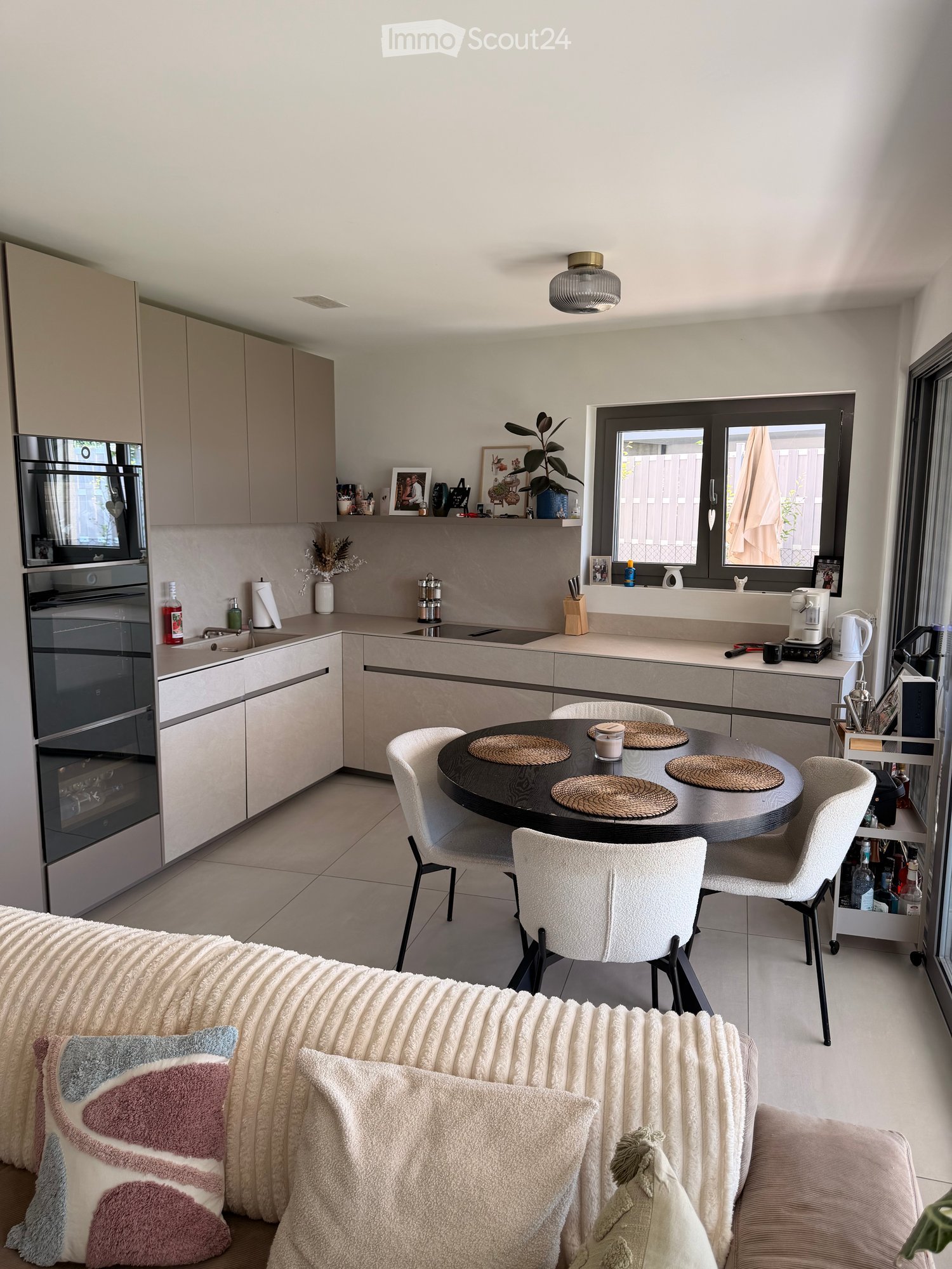 Modern kitchen and living area with a round dining table, white chairs, sofa, and various kitchen appliances.