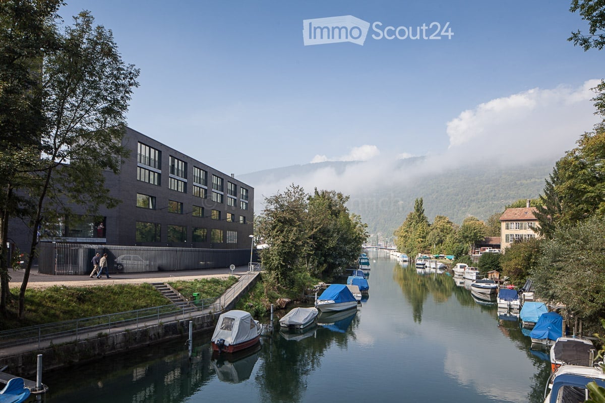 A serene canal lined with trees and docked boats, with a modern building on the left and a traditional house on the right, under a clear blue sky with distant mountains.
