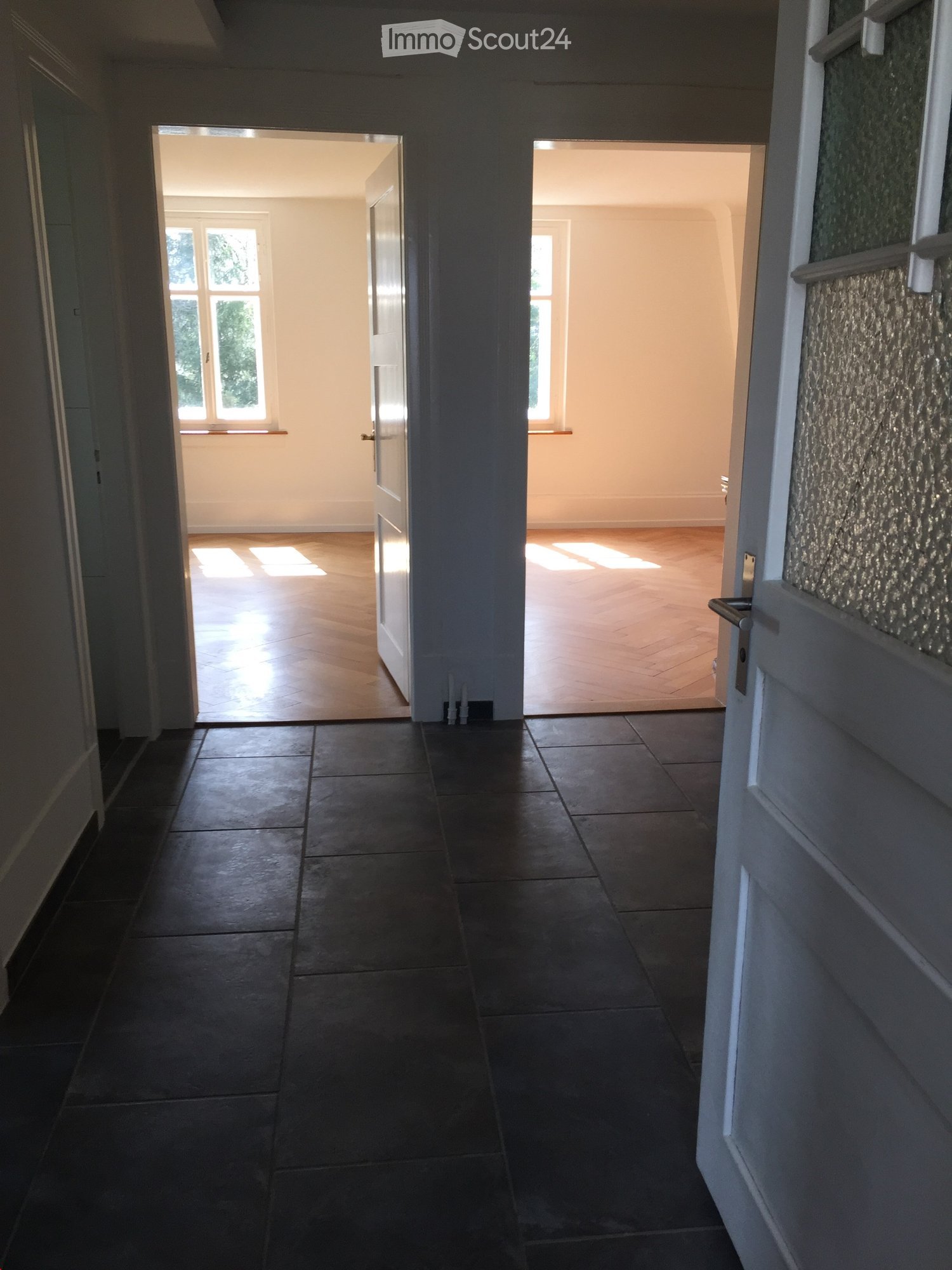 Empty hallway with dark tiled floor, light-colored walls, and two open doors leading to rooms with wooden flooring and windows.