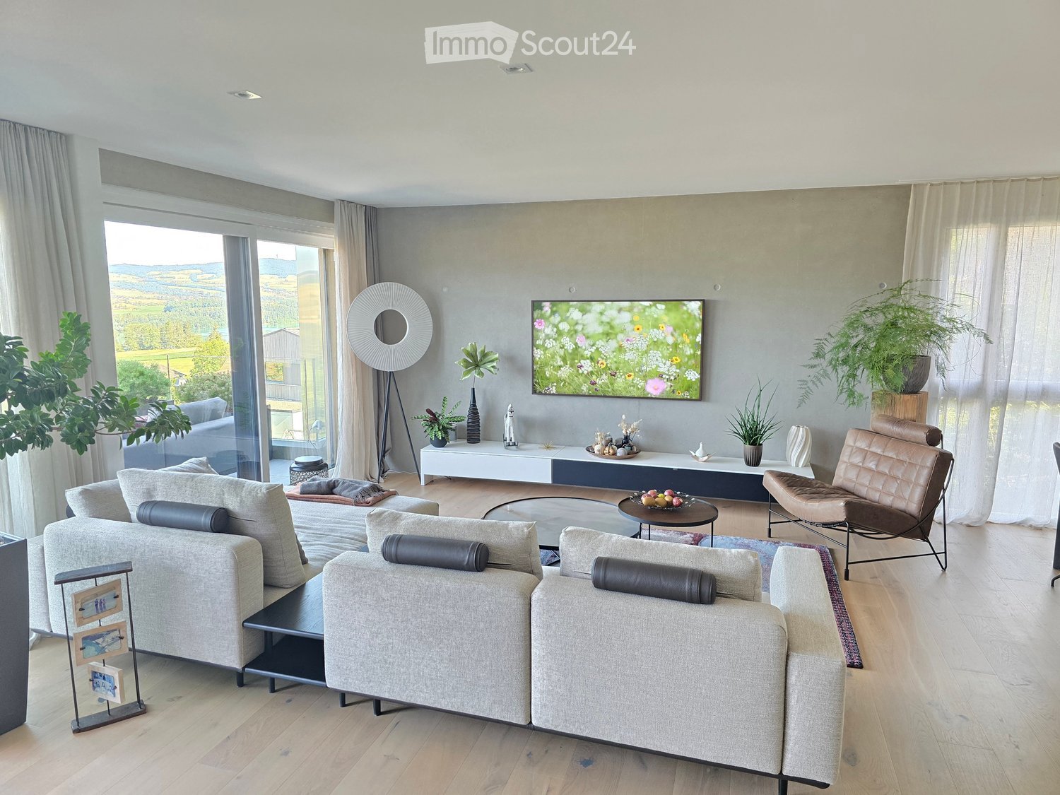 Modern living room with large windows, grey sectional sofa, coffee table, leather chair, TV, floor lamp, and decorative plants.