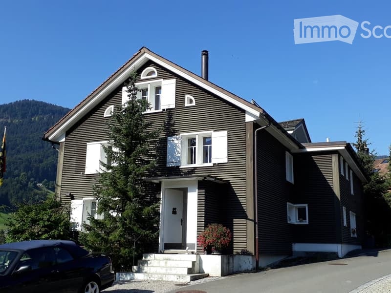 Two-story house with dark brown siding, white shutters, and a raised ground floor entrance. A small garden area with potted plants is visible near the entrance.